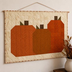 May include: A rectangular quilted wall hanging featuring three stylized pumpkins in shades of orange and brown against a cream-colored background. The piece is suspended from a wooden rod with a string and displays a grid-like stitching pattern.