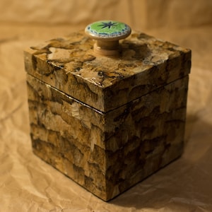 May include: A small, square wooden box with a decorative lid. The box is made of wood with a brown and tan mottled finish. The lid has a green and blue star-shaped knob.