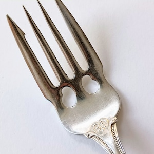 May include: Close-up of a silver fork with four tines. The fork has heart-shaped cutouts near the base and ornate detailing on the handle. The tines are sharp and pointed, and the fork appears to be made of stainless steel.