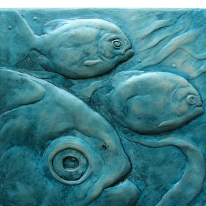 Puede incluir: Un azulejo de cerámica azul turquesa con un diseño en relieve de tres peces. Los peces están en posición de natación con sus colas y aletas visibles. El azulejo tiene una superficie texturizada y un ligero brillo metálico.