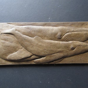 May include: A brown wooden wall plaque with a carved image of two whales, one larger than the other, swimming side by side. The whales are in a relief style, with their bodies protruding from the surface of the plaque.