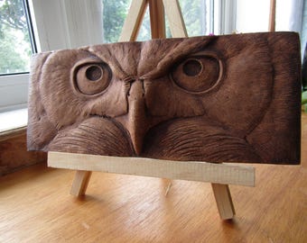 May include: A rectangular wooden plaque featuring a detailed relief carving of an owl's face. The brown wood displays intricate textures representing feathers, with deep-set eyes and a sharp beak centered on the horizontal surface.