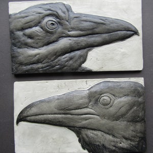 May include: Two black and white ceramic relief sculptures of crow heads. The crows are facing left and right, with their beaks open and their eyes visible.
