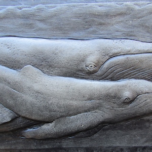 May include: A grey stone sculpture of two whales, one larger than the other, swimming side by side. The whales are carved in detail, with visible eyes, mouths, and fins.