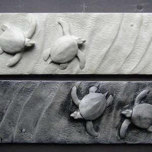 May include: Two rectangular wood carvings of sea turtles swimming in the ocean. The top carving is painted white and the bottom carving is painted black.