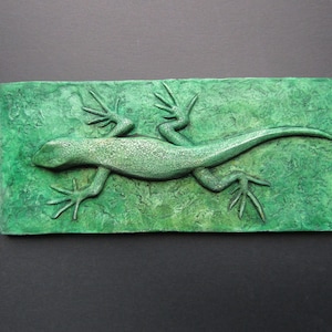 Puede incluir: Una escultura de lagarto verde sobre un fondo verde texturizado. El lagarto está en una pose de carrera con las patas extendidas.