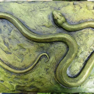 May include: A green snake sculpture with a textured surface, set against a green background. The snake is coiled in a circular shape with its head raised.