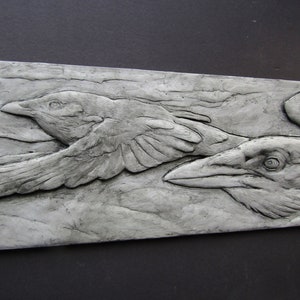 May include: A gray, textured, rectangular wall sculpture featuring two stylized crows in flight. The crows are carved in relief and appear to be flying in opposite directions.