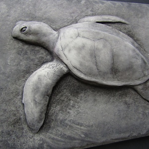 Peut inclure: Une tortue de mer sculptée en gris avec une carapace et des nageoires détaillées. La tortue est montée sur un fond gris texturé.