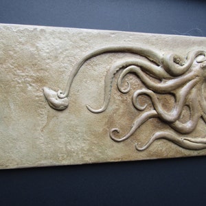 Puede incluir: Una placa de pared de cerámica beige con una escultura en relieve de un pulpo con ocho tentáculos. El pulpo está enroscado con sus tentáculos extendiéndose en diferentes direcciones.