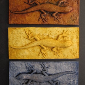 May include: Three ceramic wall plaques, each featuring a stylized lizard in a different color: brown, yellow, and blue. The lizards are sculpted in a 3D relief style, giving them a textured appearance.