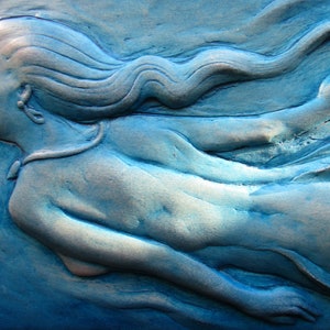 May include: A blue ceramic sculpture of a woman with flowing hair, sculpted in a wave-like pattern. The sculpture is a bas-relief, meaning it is raised from the background.