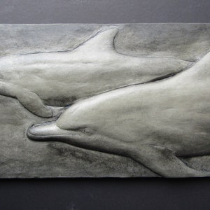 May include: A gray and white ceramic wall sculpture of two dolphins swimming side by side.