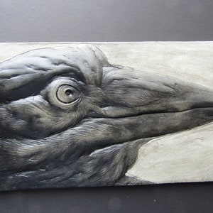 May include: A black and white ceramic relief sculpture of a crow's head. The crow's eye is detailed and the feathers are textured.