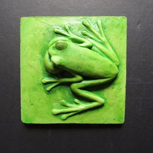 Puede incluir: Una escultura de pared de cerámica verde de una rana con una superficie texturizada. La escultura está firmada "HARE" en la esquina inferior derecha.