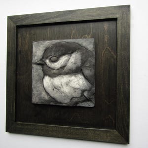 May include: A square wall art piece featuring a relief sculpture of a small bird with closed eyes, rendered in shades of gray and white. The sculpture is mounted on a dark, stained wooden square panel and enclosed within a matching dark wood frame.