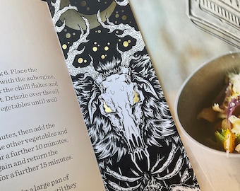 May include: A black and white bookmark with a gold foil design. The bookmark features a skull with glowing eyes and a crescent moon in the background. The skull is surrounded by branches and leaves.
