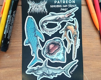 May include: A black sticker sheet with illustrations of different sharks, such as a whale shark and a hammerhead shark. The sheet includes the text "PHASEMOTH" and "PATREON NOVEMBER 2024 STICKER SHEET". The stickers are surrounded by pens.