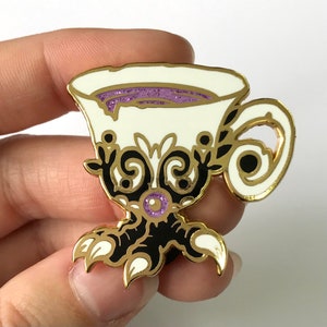 May include: A gold and white enamel pin of a teacup with a purple glitter interior and black claws. The teacup has a gold rim and intricate black and gold designs.