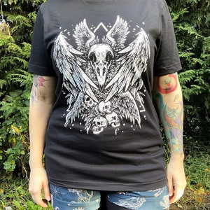 Peut inclure: T-shirt noir avec un imprim&eacute; blanc d'un cr&acirc;ne avec des ailes et une cha&icirc;ne de cr&acirc;nes. Le motif est centr&eacute; sur le t-shirt.