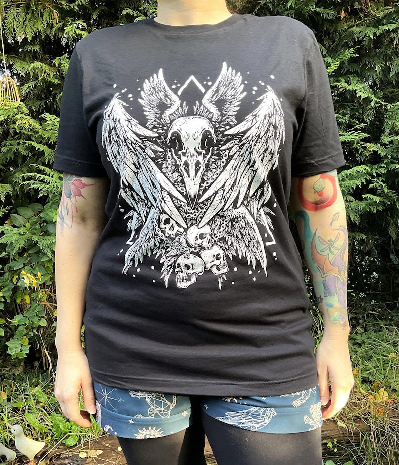 Peut inclure: T-shirt noir avec un imprim&eacute; blanc d'un cr&acirc;ne avec des ailes et une cha&icirc;ne de cr&acirc;nes. Le motif est centr&eacute; sur le t-shirt.