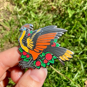 May include: A colorful enamel pin featuring a stylized bird with orange, yellow, black, and gray feathers. The bird is perched on a bed of green leaves and red berries. The pin has a silver outline.