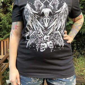 Peut inclure: T-shirt noir avec un imprim&eacute; graphique blanc d'un cr&acirc;ne avec des ailes et des cha&icirc;nes. Le cr&acirc;ne est entour&eacute; de trois petits cr&acirc;nes.