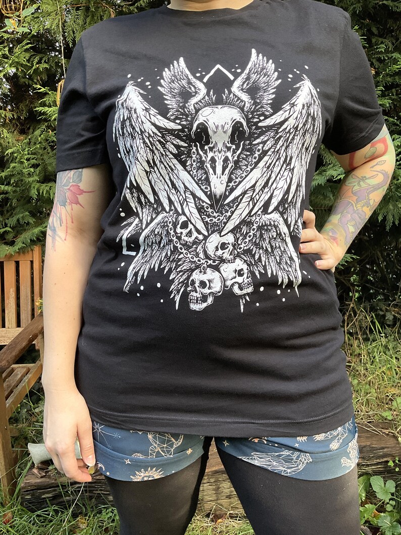 Peut inclure: T-shirt noir avec un imprim&eacute; graphique blanc d'un cr&acirc;ne avec des ailes et des cha&icirc;nes. Le cr&acirc;ne est entour&eacute; de trois petits cr&acirc;nes.