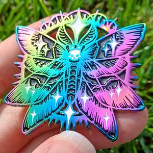 May include: A vibrant moth-shaped pin badge with a skull motif. The pin showcases a rainbow gradient of colours, including blue, green, and pink, with black detailing and white star accents. The moth has intricate wing patterns and a circular border.