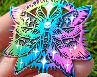 May include: A vibrant moth-shaped pin badge with a skull motif. The pin showcases a rainbow gradient of colours, including blue, green, and pink, with black detailing and white star accents. The moth has intricate wing patterns and a circular border.
