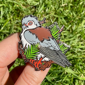 Könnte beinhalten: Eine silberne Emaille-Anstecknadel mit einem grau-braunen Vogel mit roten Augen, der auf einem grünen, belaubten Zweig sitzt.