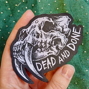 May include: A black and white patch with a detailed illustration of a saber-toothed tiger skull. The text "DEAD AND DONE" is printed in white, gothic-style lettering. The patch is held against a green background.