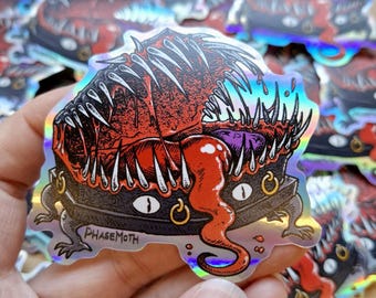 May include: A holographic sticker featuring a dark, toothy creature with a wide, open mouth, sharp white fangs, and a long, red, dripping tongue. The creature has multiple eyes and small clawed feet, with the text PHASEMOTH printed on the lower left side.