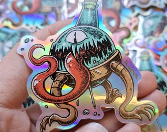 May include: A holographic sticker featuring a round, teal creature with a single large eye, sharp teeth, and a long red tongue protruding from a glass bottle top. The creature has tan, segmented limbs and the text PHASEMOTH printed on the side.