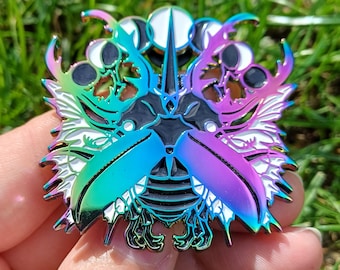 May include: An iridescent metal pin with a detailed insect design. The pin features a central black shape with a multi-coloured beetle-like form, with blue, green, purple, and white accents. The pin is held against a background of green grass.