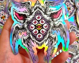 May include: Holographic sticker depicting a fantastical creature with multiple eyes, wings, and a furry body. The sticker has a rainbow sheen and is outlined in black. The creature has pink ears and a central eye cluster. The text "PHASEMOTH" is printed on the sticker.
