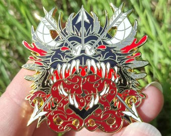 May include: A metal enamel pin featuring a stylized creature head with sharp white teeth, a red tongue, and dark blue accents. The design includes two crossed white arrows behind the head, with gold metal outlines and red decorative elements.