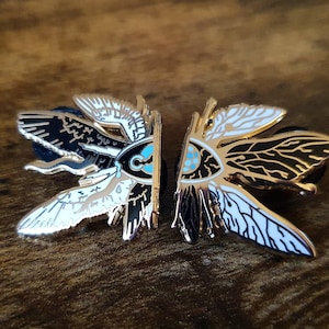 May include: Two matching moth-shaped enamel pins. Each pin features a gold outline with black, white, and teal details. The pins are symmetrical, with one side black and the other white, creating a striking contrast. The pins are on a wooden surface.