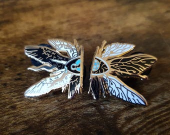 May include: A pair of moth-shaped enamel pins. Each pin has a gold outline with black, white, and teal details. The pins are mirror images of each other, with one side black and the other white, creating a contrast. The pins are on a wooden surface.