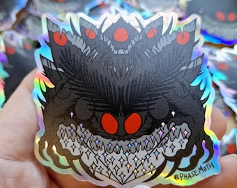 May include: A holographic sticker featuring a stylised black moth creature with large, glowing red eyes, a jagged mouth filled with sparkling diamond-shaped teeth, and the text @PHASEMOTH printed in the bottom right corner of the design.