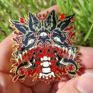 Puede incluir: Un pin de esmalte metálico que presenta una cabeza de lobo negra estilizada con un tercer ojo, dientes blancos afilados y una lengua azul larga. El diseño incluye una columna vertebral blanca central rodeada de salpicaduras rojas y bordes dorados.