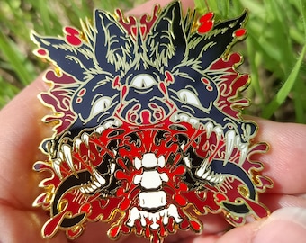 May include: A metal enamel pin featuring a stylized black wolf head with a third eye, sharp white teeth, and a long blue tongue. The design includes a central white spinal column graphic surrounded by vibrant red splatters and gold metal outlines.