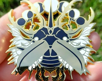 May include: A gold-coloured metal pin with a detailed design of a stylised insect. The pin features a combination of gold, white, and navy blue enamel, with intricate wing and antenna details. The pin is held in a person's hand against a blurred green background.