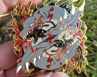 May include: A circular enamel pin featuring a stylized gray shark with sharp white teeth, surrounded by red coral-like accents and black fish silhouettes. The design includes gold metal outlines and a circular gray background, creating a detailed graphic illustration.