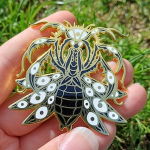 Peut inclure: Épingle en métal doré en forme d'insecte stylisé avec de multiples yeux. L'épingle présente des détails en émail noir et blanc, avec un motif détaillé sur les ailes et le corps. L'épingle est tenue sur un fond d'herbe verte.