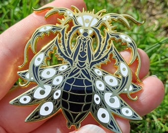 May include: A gold-coloured metal pin in the shape of a stylised insect with multiple eyes. The pin features black and white enamel details, with a detailed design on the wings and body. The pin is held against a background of green grass.