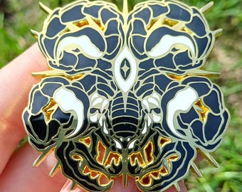 May include: An enamel pin with a scorpion design. The pin features a black and white colour scheme with gold accents. The scorpion's body is black, with white claw-like shapes and a gold outline.