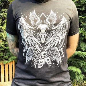 Peut inclure: T-shirt noir avec un imprim&eacute; graphique blanc d'un cr&acirc;ne avec des ailes et des cha&icirc;nes. Le cr&acirc;ne est entour&eacute; de plumes et de cr&acirc;nes.