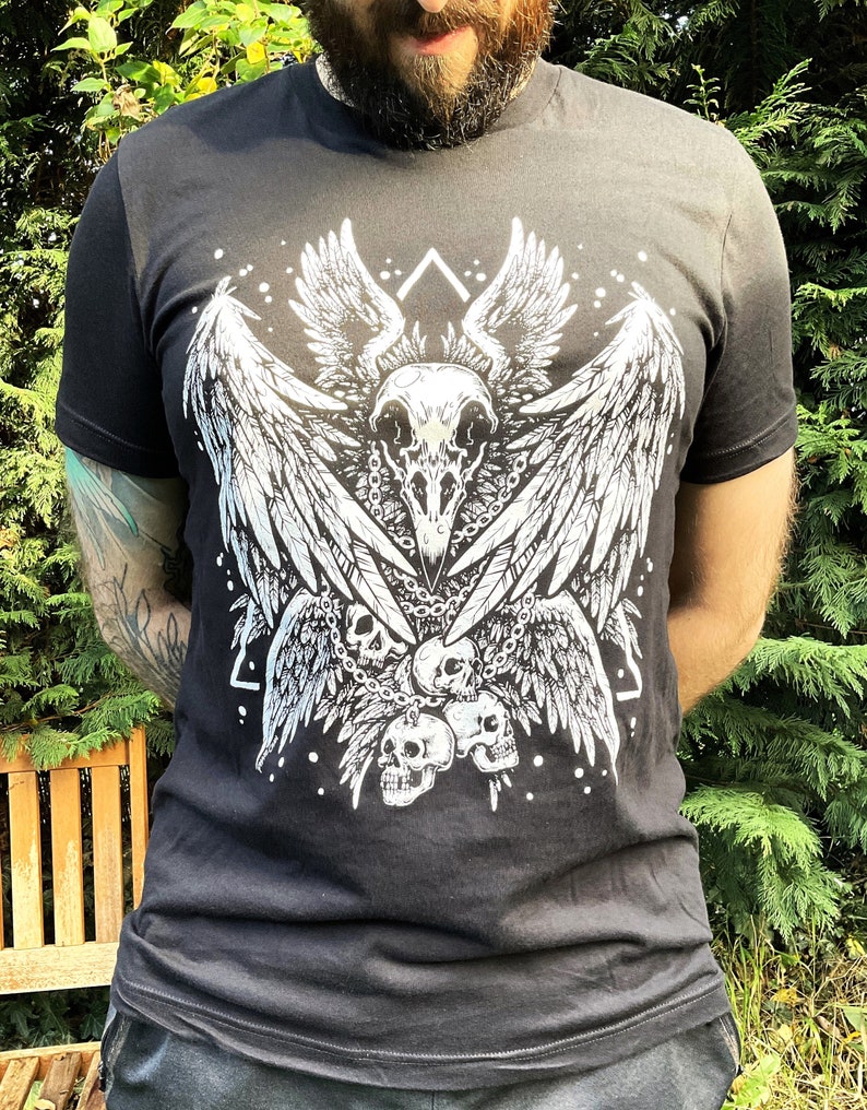 Peut inclure: T-shirt noir avec un imprim&eacute; graphique blanc d'un cr&acirc;ne avec des ailes et des cha&icirc;nes. Le cr&acirc;ne est entour&eacute; de plumes et de cr&acirc;nes.