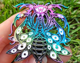 May include: An iridescent insect pin with a detailed design. The pin features a stylized insect with multiple eye-like shapes and a gradient of colours, including blue, pink, and green. The pin is made of metal and has a glossy finish.